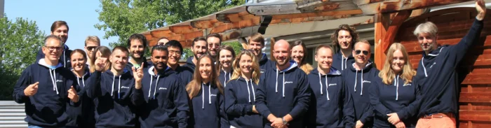 Group photo of the Picterra team outdoors wearing company hoodies, smiling together in front of a wooden structure