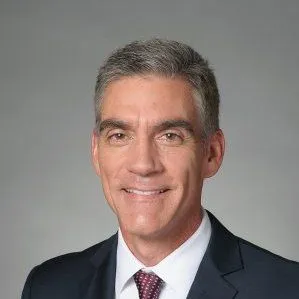Professional headshot of a man in a dark suit and tie smiling at the camera against a gray background