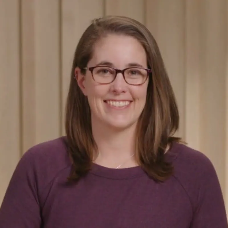 Professional headshot of a woman wearing glasses and a purple sweater, smiling at the camera.