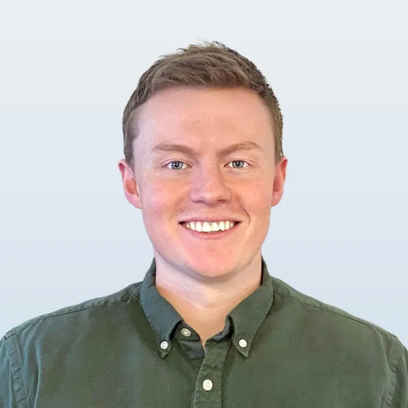 A smiling man with short light-brown hair wearing a green button-up shirt stands in front of a pale, neutral background.